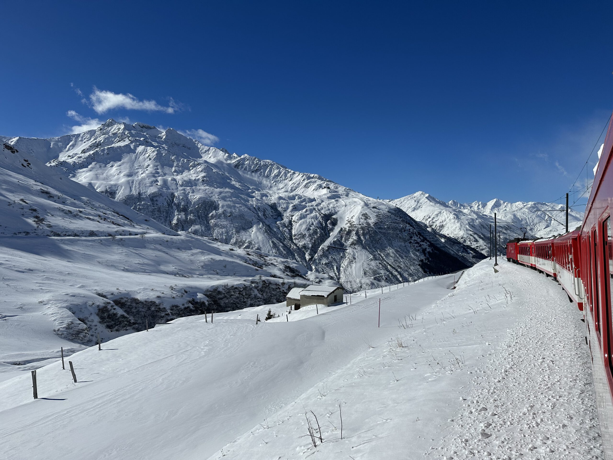 2023 Andermatt/Oberalp-Tschamut: Bahnstrecke mit roten Leitplanken durch tiefen Schnee, dahinter Berge