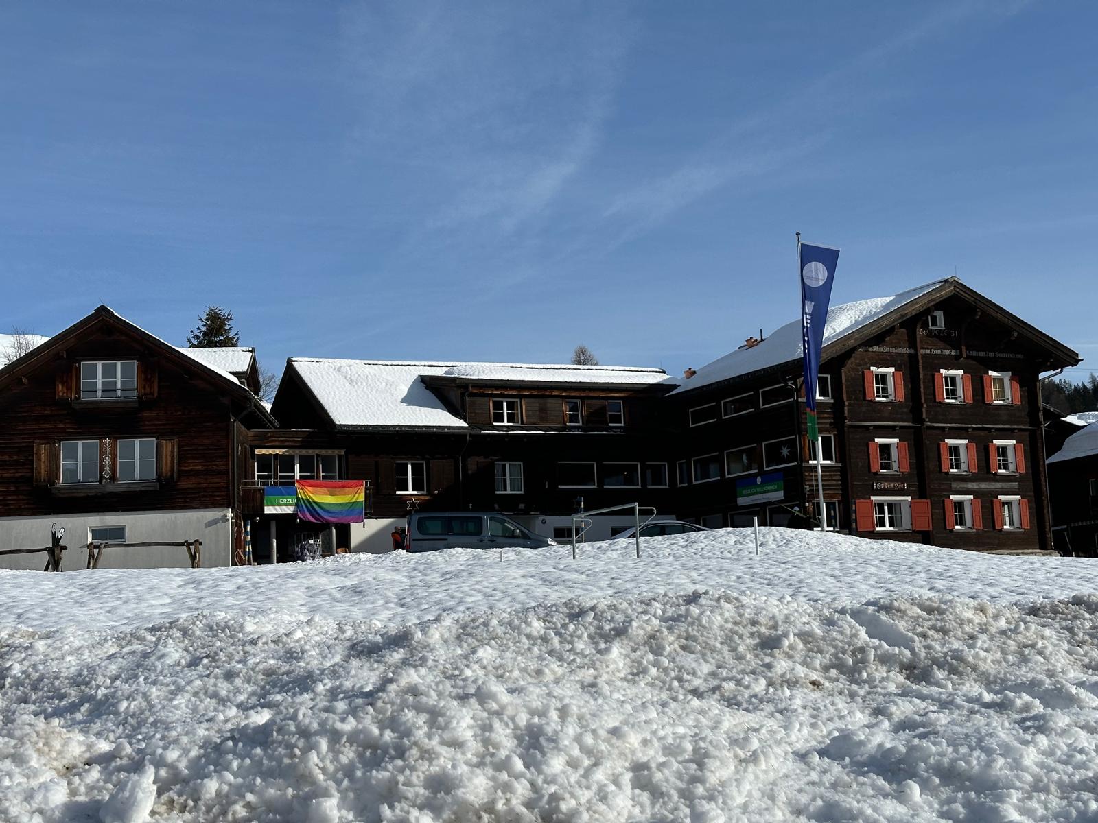 2024 Davos: Dunkles Holzhaus mit Fahne im Schnee, dahinter der Hang