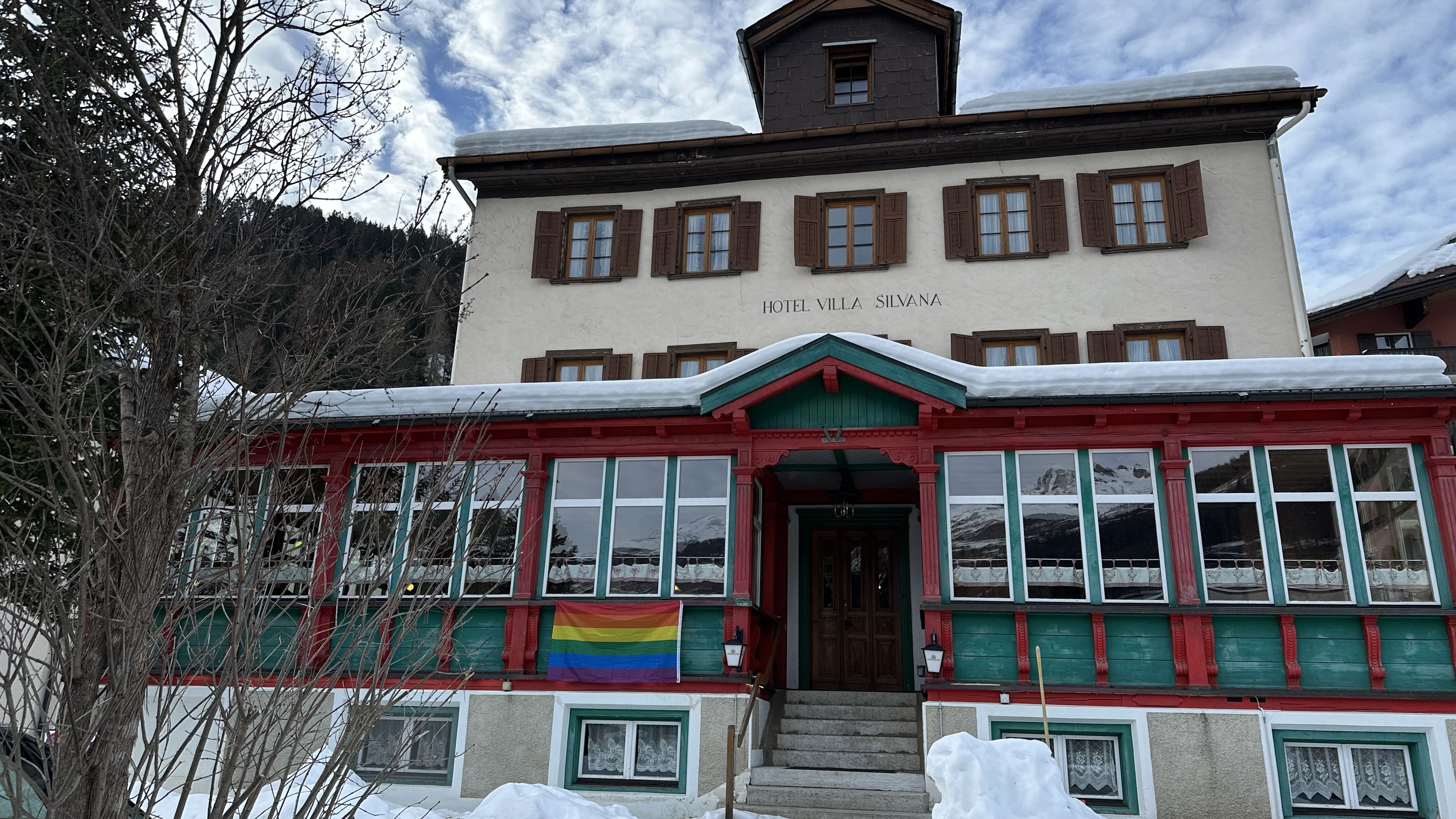 2025 Scuol: Das Gruppenhaus von vorn, Riegelbau mit grün und rot gefasster Veranda, davor Schnee