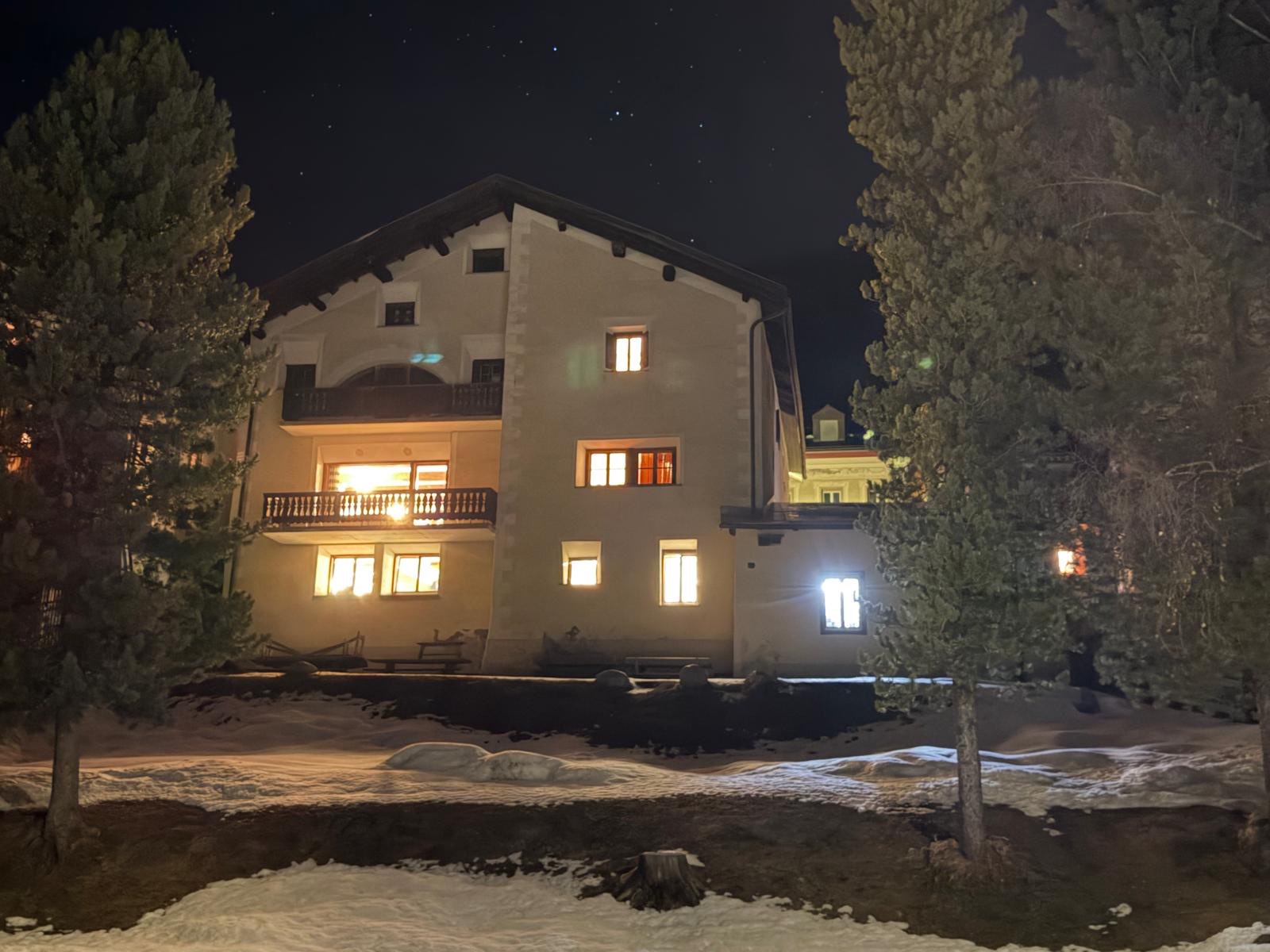 2026 S-chanf: Grosses Chalet bei Nacht, warm erleuchtete Fenster, davor Schnee und Tannen